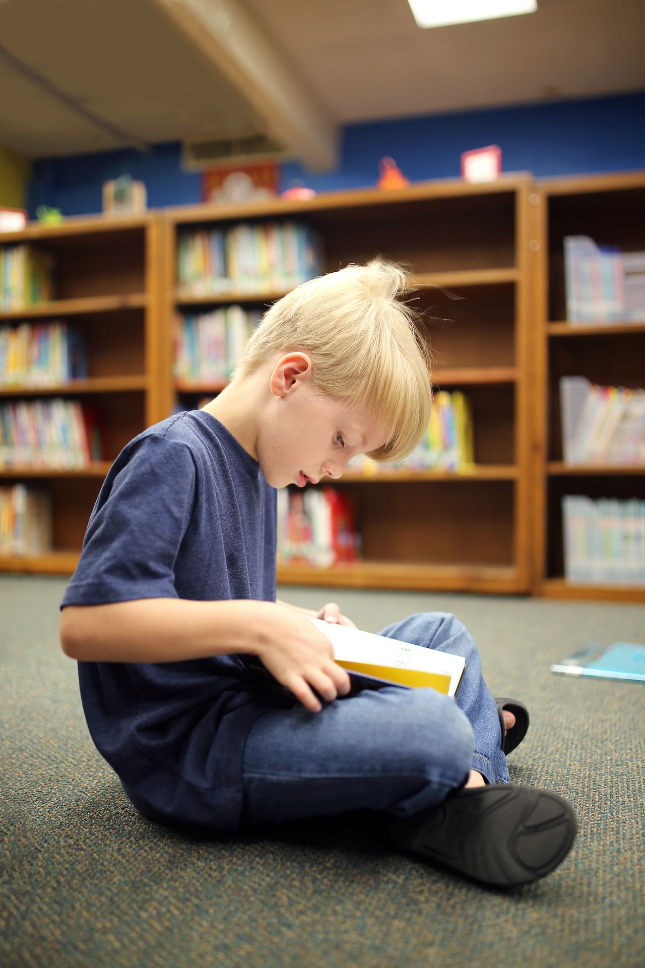 A boy sits cross-legged on a library floor reading a book in front of full shelves.