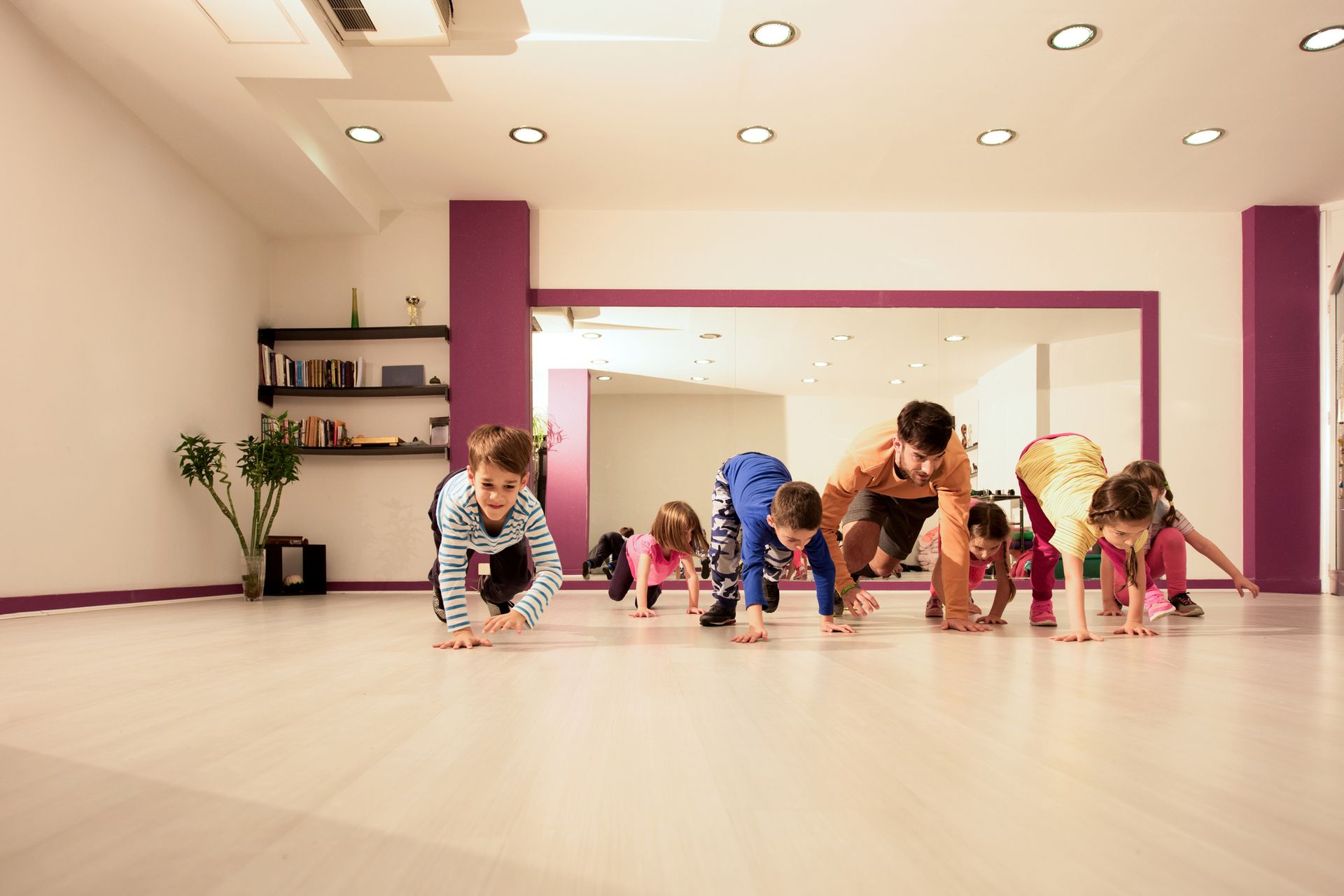 Young children crawl across a wooden studio floor on hands and feet as an adult leads them.