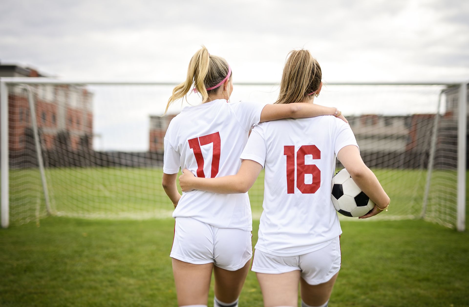 Two soccer players in white jerseys walk arm in arm toward a goal, one carrying a ball.