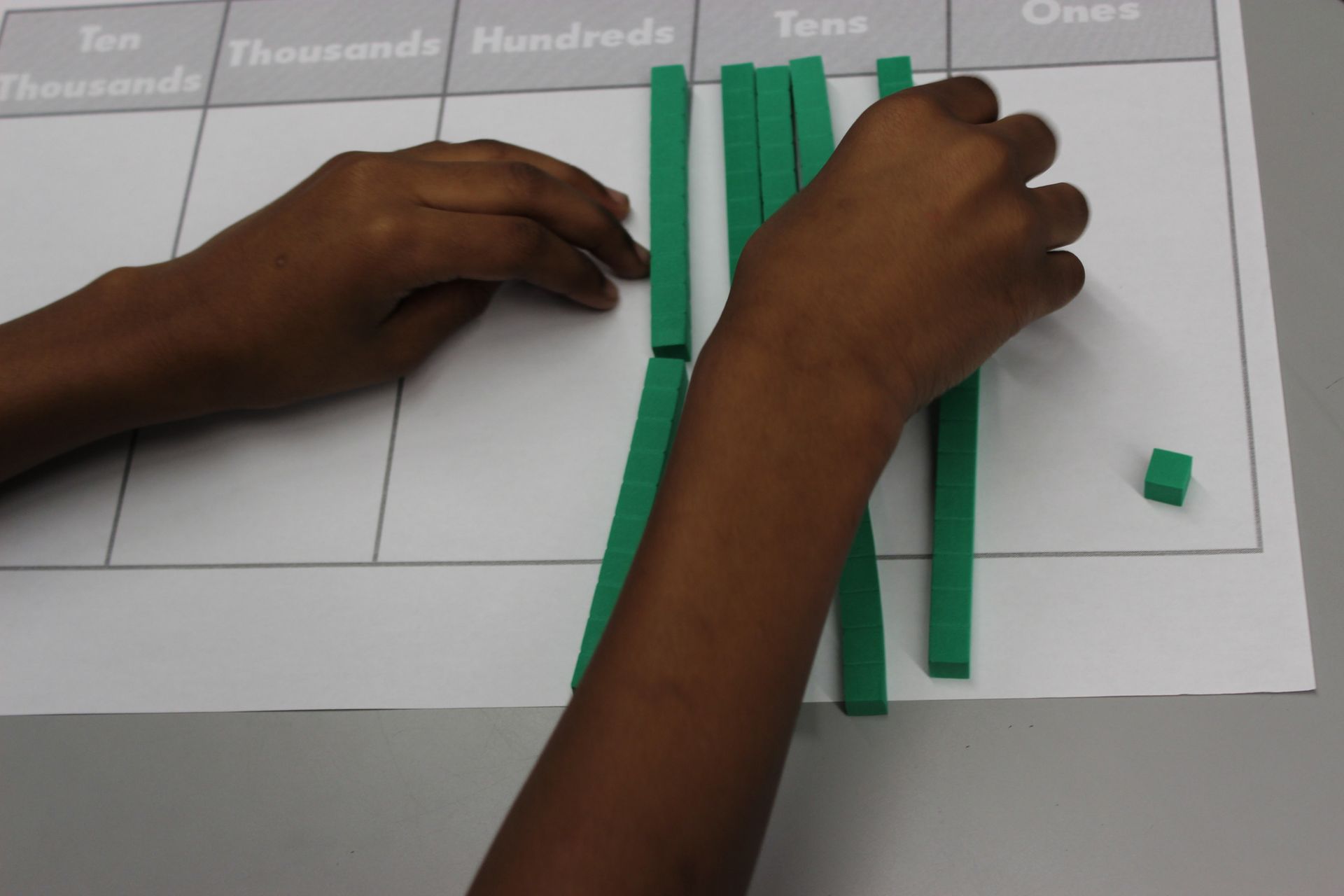 Hands line up green counting rods on a place value chart marked tens and ones.